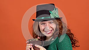 Young woman celebrating saint patrick`s day on orange wall in hat holding pot with money