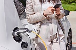 Young woman charging an electric car at public charging station and pays using a mobile phone. Innovative eco-friendly vehicle.