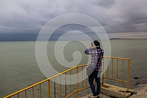 Young woman in checkered shirt standing on the beach alone and looking at sea in a gloomy gray day. Back view