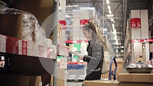 Young woman is checking her list, taking a sink and pipes from a shelf and putting them on the trolley in a warehouse