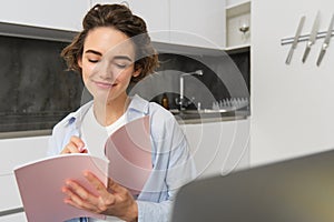 Young woman checking her schedule while working on remote from home, using laptop, looking at her daily planner, making