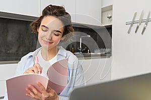 Young woman checking her schedule while working on remote from home, using laptop, looking at her daily planner, making