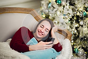 Young woman checking out her text messages on mobile phone as she liyng sofa in front of the Christmas tree at home.