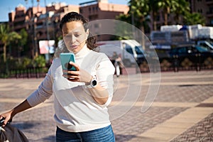 Young woman checking smart phone on the street, booking taxi or searching geolocation on online maps on mobile app