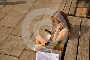 Young woman checking social networks on her cell phone sitting on a bench surrounded by shopping bags seen from above