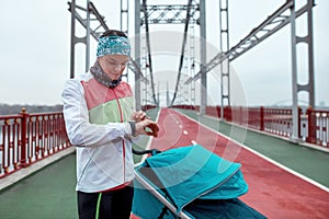 Young woman checking time and results using smart watch on her wrist while standing on the bridge with a baby carriage