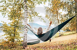 The young woman cheerfully rose arms up while she swinging in a hammock between the birch trees on the mountain lake bank. Out-of-