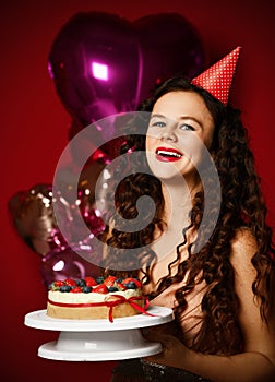 Young woman chef cook holding sweet cake with strawberry blueberry and cream smiling on dark red background