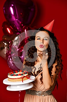 Young woman chef cook holding sweet cake with strawberry blueberry and cream smiling on dark red background