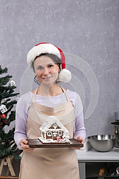 Young woman chef in New Years hat holds gingerbread house in her hands. Vertical frame