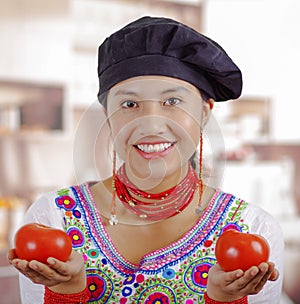 Young woman chef wearing traditional andean blouse, black cooking hat, holding tomatoes up showing to camera and smiling