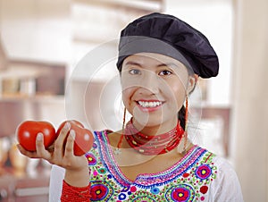 Young woman chef wearing traditional andean blouse, black cooking hat, holding tomatoes up showing to camera and smiling