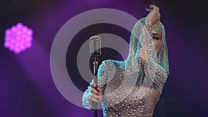 A young woman in a chic, tight-fitting jumpsuit with rhinestones and a shiny headdress dances near a vintage microphone