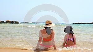 Young woman and child girl sit with their backs to camera, on a sandy beach, chatting, enjoying warm water of the sea