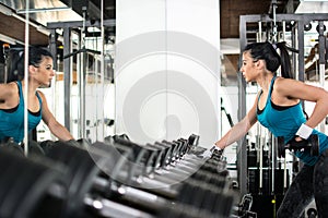 Young woman choosing dumbbells in gym. She stay near a mirror and see her reflection.