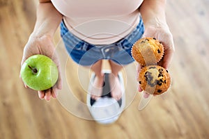 Young woman choosing between fruit and sweet on weighing scale at home. Diet concept.