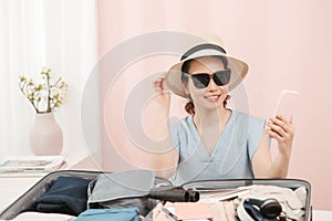 Young woman choosing hat sitting with clothes and suitcase on the bed. Prepairing for the summer vacations