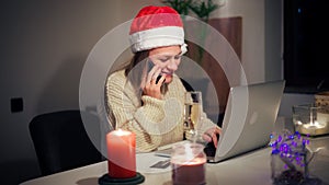 Young woman in a Christmas hat sits in front of a laptop and speaks on the phone