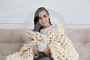 Young woman with chunky knit blanket and cup on sofa at home