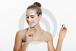 Young woman with clay facial mask holding cucumber slices isolated on white background