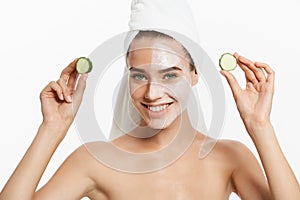 Young woman with clay facial mask holding cucumber slices on white background