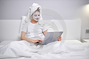 Young woman with clay facial mask using laptop while sitting on bed at home.