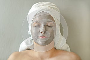 Young woman with clay mask on face and towel on head in form of turban lies with closed eyes and enjoys spa treatment