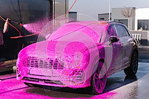 Young woman cleaning her car on self service car wash. Female spread soap using jet sprayer. Pink foam on the car