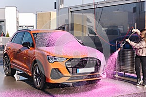 Young woman cleaning her car on self service car wash. Female spread soap using jet sprayer. Pink foam on the car