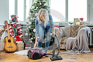 Young woman cleaning with vacuum cleaner, vacuuming under Christmas Tree needles with New Years ornaments on hardwood