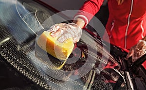 Young woman cleaning windshield of her car, closeup detail on hand in glove holding yellow sponge