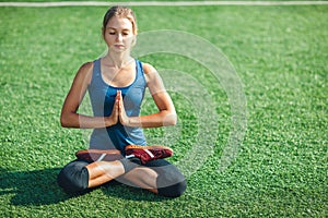 Young woman with closed eyes in a blue shirt and leggings enjoying meditation and yoga on green grass in the summer on nature.