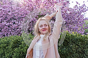 Young woman with closed eyes smiling and stand in blooming sakura park raised her hands. Happy woman in spring park