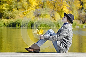 Young woman with closing eyes sitting on the pier, autumn leaves