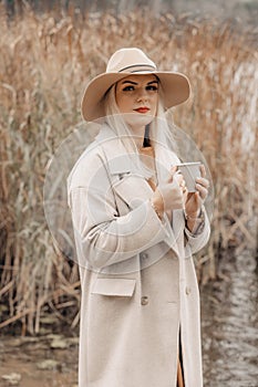 A young woman in a coat and hat enjoy nature and a cup of hot tea on the shore of a lake