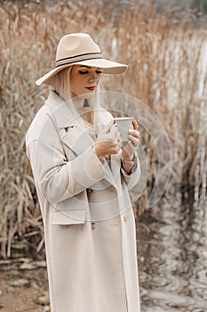 A young woman in a coat and hat enjoy nature and a cup of hot tea on the shore of a lake