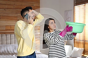 Young woman collecting leaking water from ceiling while her boyfriend calling roof repair service at home