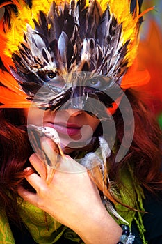 Young woman with a colorful feather carnival face mask on bright colorful background, eye contact, make up artist.