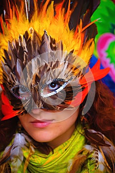 Young woman with a colorful feather carnival face mask on bright colorful background, eye contact, make up artist.