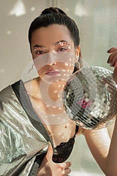 Young woman with colorfull spice makeup and Mirror disco ball with shines on shes body near window