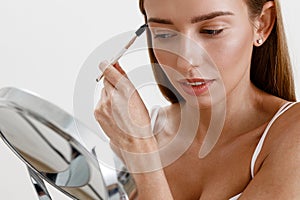 Pretty woman combing her eyebrows with brush and looking at mirror over white studio background