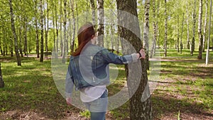 Young woman comes up to tree puts hand on it, turns around her head and smiles looking at camera