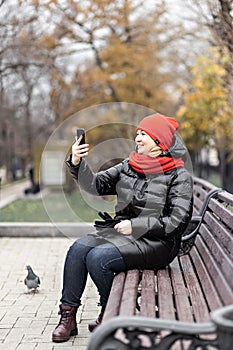 A young woman communicates in online video chat on a mobile phone, sitting on a bench in an autumn park. Social distancing and