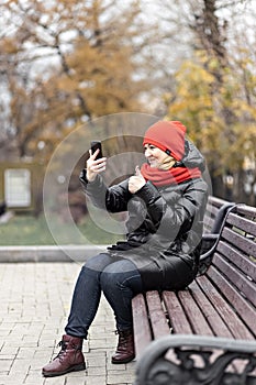 A young woman communicates in online video chat on a mobile phone, sitting on a bench in an autumn park. Social distancing and