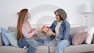 Young woman congratulating her husband with present and embrace him, sitting on couch at home, slow motion