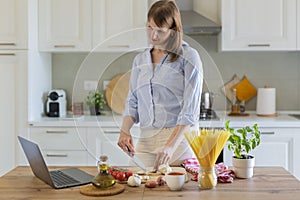 A young woman cooks at home in the kitchen, she watches a video recipe on a laptop and works with the dough on the table, she cuts