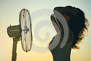 Young Woman cooling herself during hot weather in front of fan on a blue sunset sky background