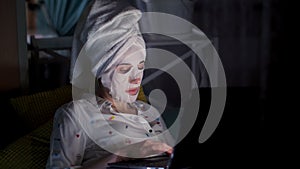 Young woman in cosmetic fabric mask and white towel on her head, is working on laptop, watching on screen, at night