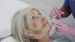 Young woman cosmetologist in uniform making injections for lip augmentation and anti-wrinkle of elderly female in