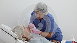 Young woman cosmetologist in uniform making injections for lip augmentation and anti-wrinkle of elderly female in
